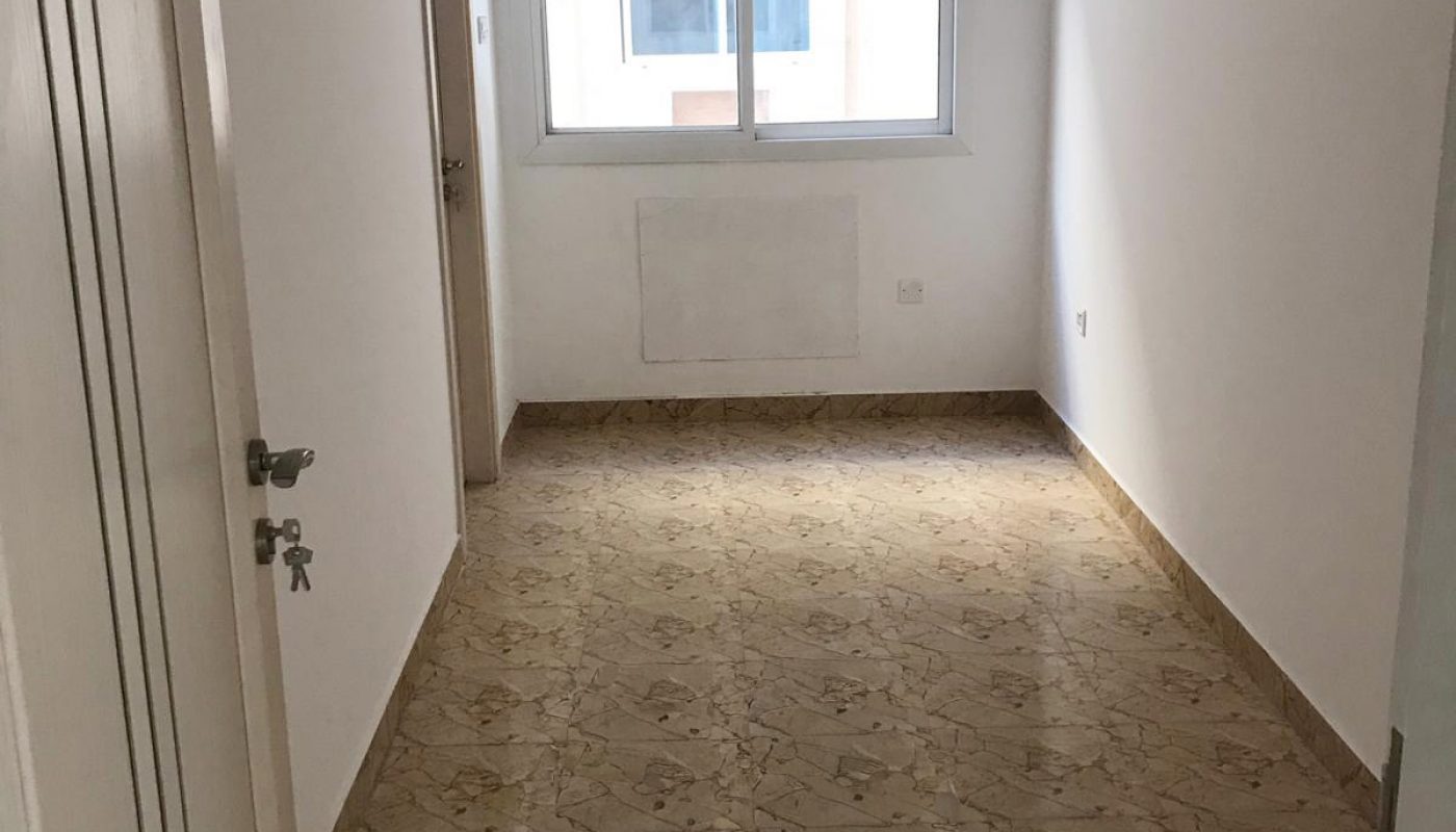 Narrow, empty room with tiled floor, white walls, ceiling fan, closed window, and a wall-mounted light fixture.