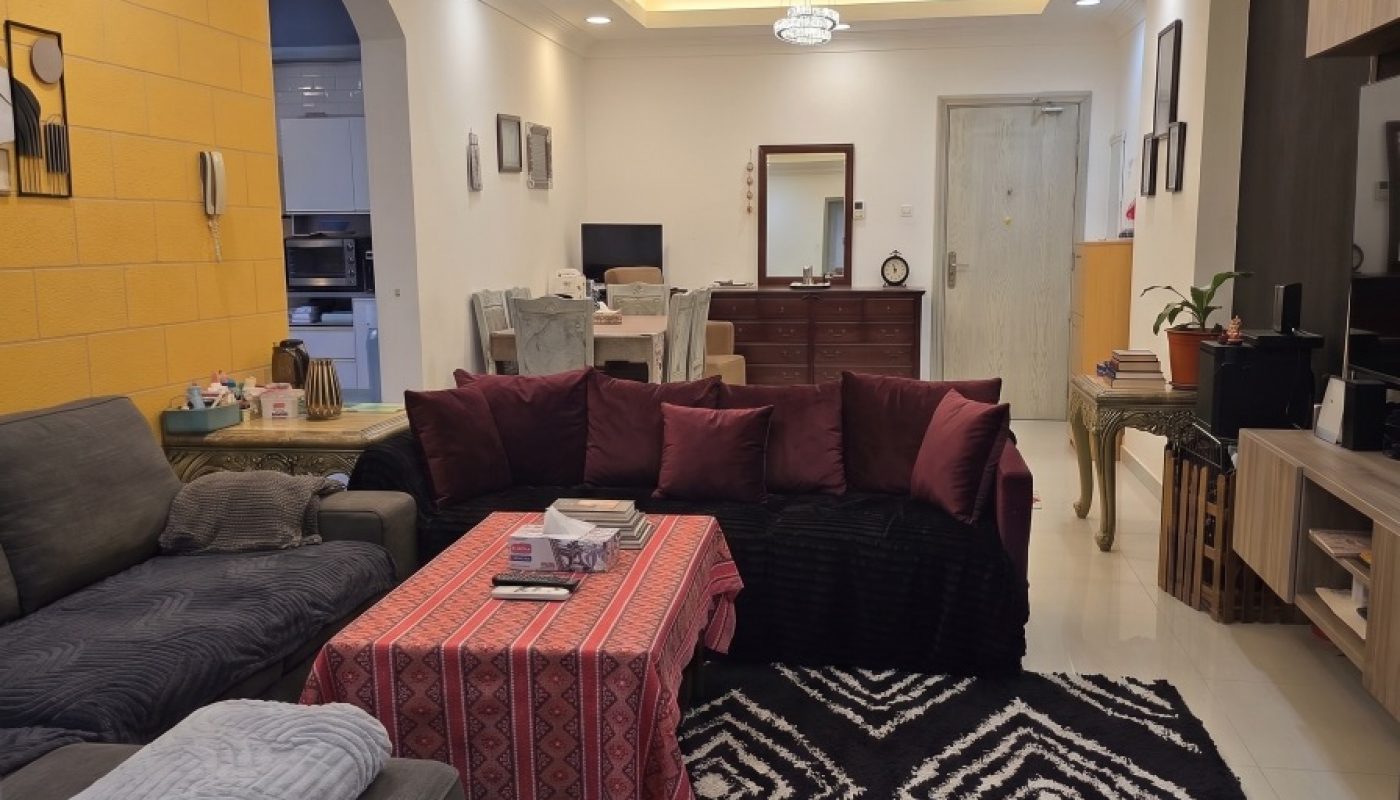 A modern living room with gray sofas, a red tablecloth, patterned rug, wall-mounted shelves, and a dining area in the background. Warm ceiling lights illuminate the space.