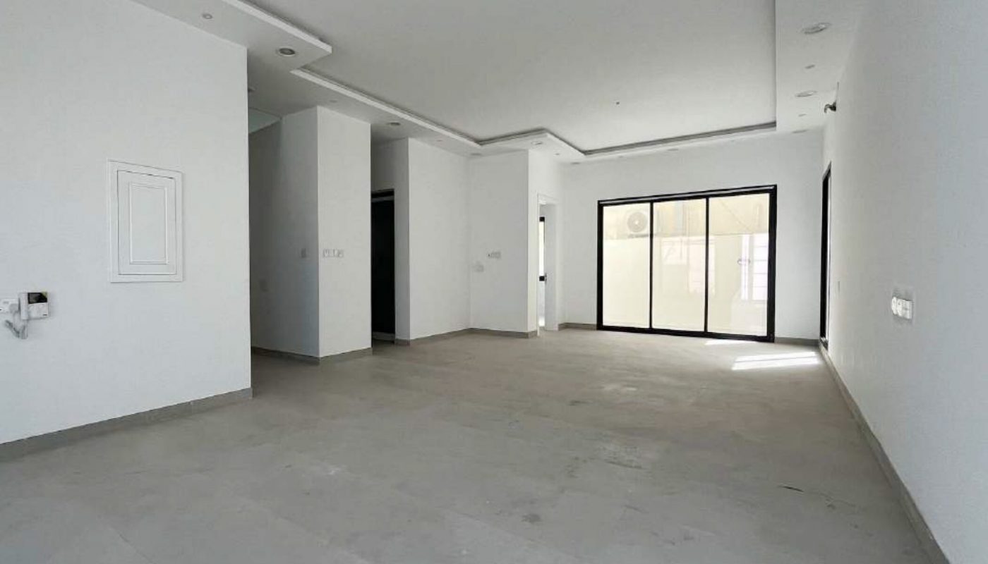 Empty, unfurnished room with white walls, large windows at the far end, recessed ceiling lights, and plain gray flooring. Natural light enters through the windows.