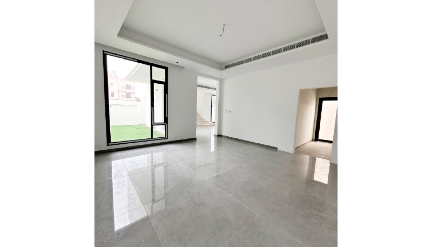 Spacious, empty room with large grey tiled floor, white walls, recessed ceiling, and windows letting in natural light; a doorway leads to another area.