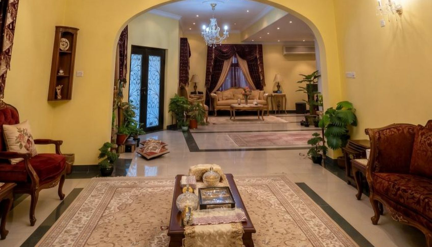 A well-lit, elegant living room with yellow walls, chandeliers, ornate furniture, potted plants, and a patterned rug, leading to a similarly styled sitting area.