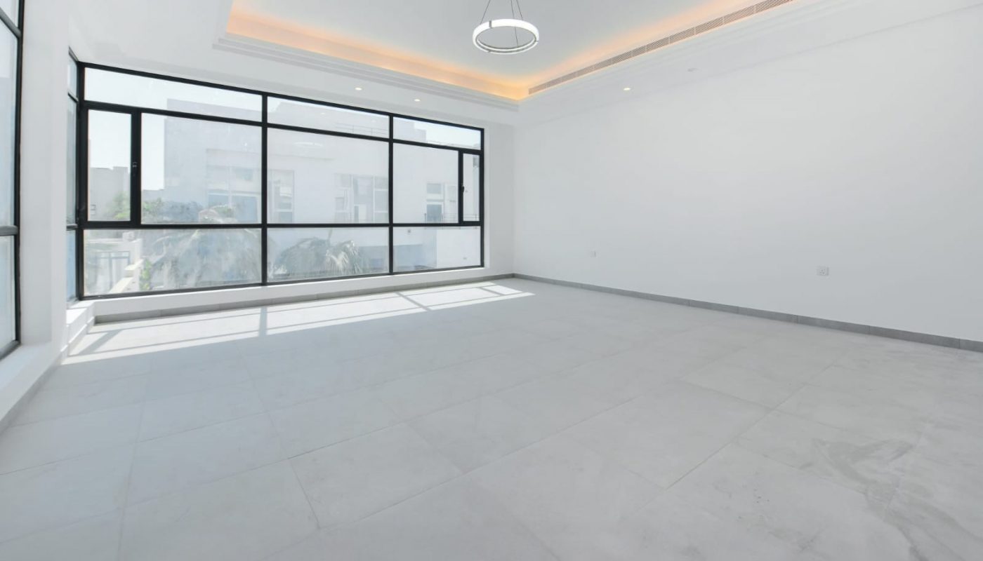 Spacious empty room with large windows, white walls, recessed ceiling lights, and a modern circular pendant light. Natural light fills the room, highlighting the tiled floor.