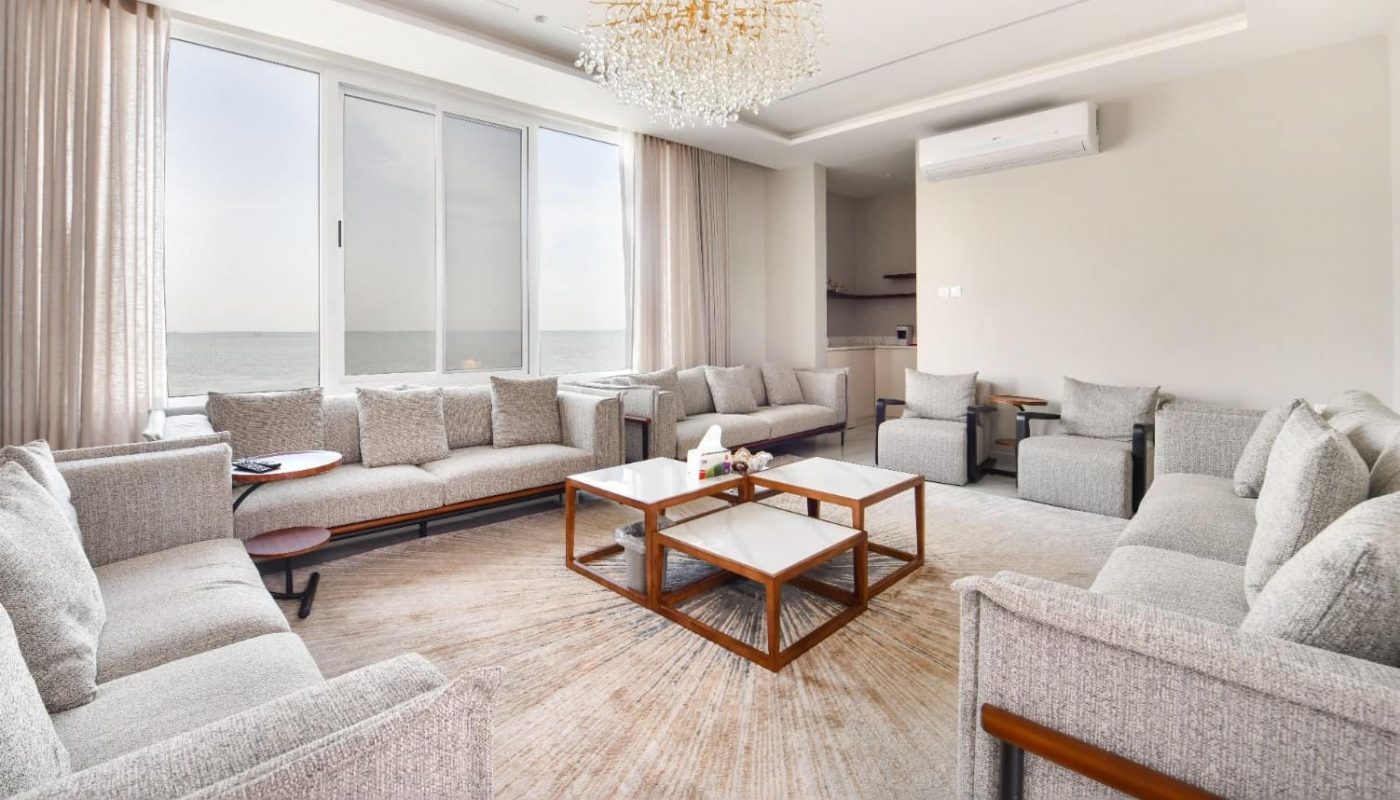 Spacious living room with large windows, neutral-colored sofas, a wooden coffee table, and a crystal chandelier. The ocean is visible outside the window.