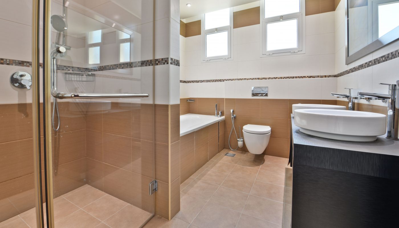 Luxury villa bathroom interior with a glass shower, bathtub, toilet, and sink with wood paneling and tiled walls.