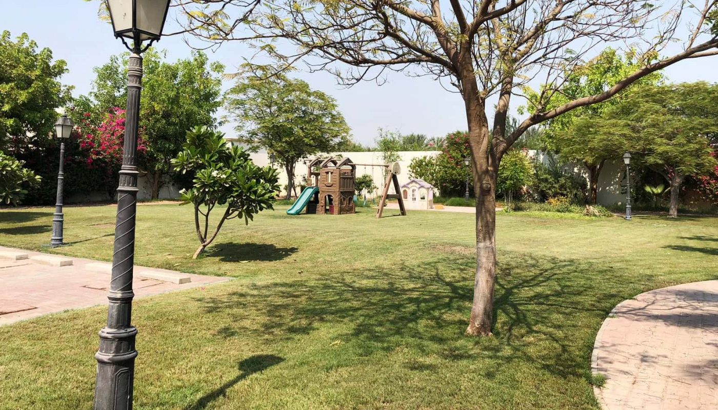 A park with green grass, trees, lamp posts, and a children's play area featuring a playhouse and a slide enhances the ambiance of the SF 4BR Villa for rent in Hamala. A winding pathway is also visible on the right side.