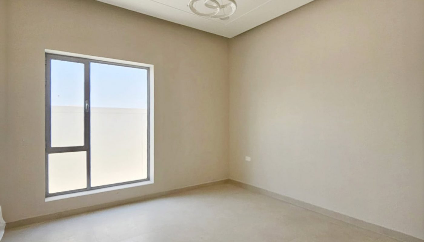 Empty room with beige walls, a large window, and a ceiling light fixture. The floor is tiled, and natural light enters from the window.