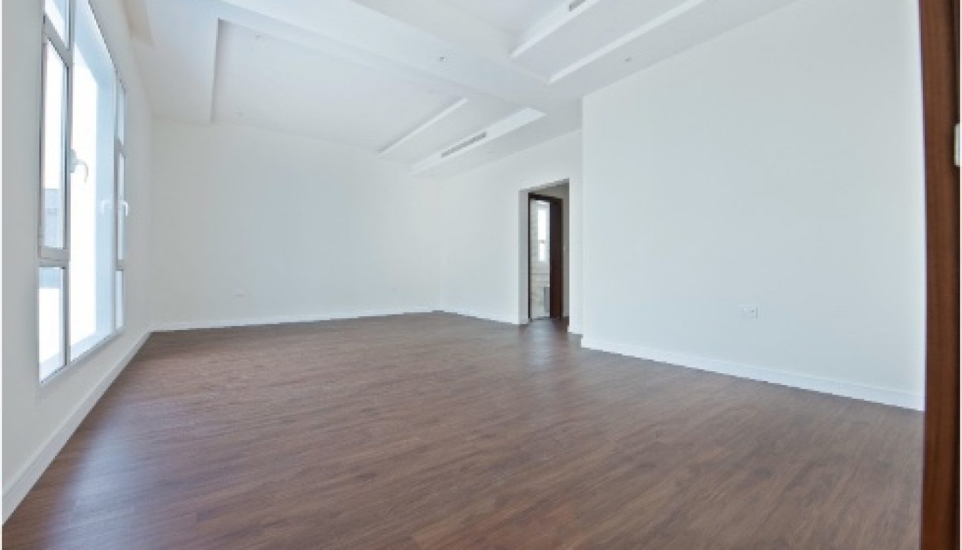 Spacious empty room with white walls, large windows, and dark wooden floor, featuring a modern coffered ceiling and an open doorway.