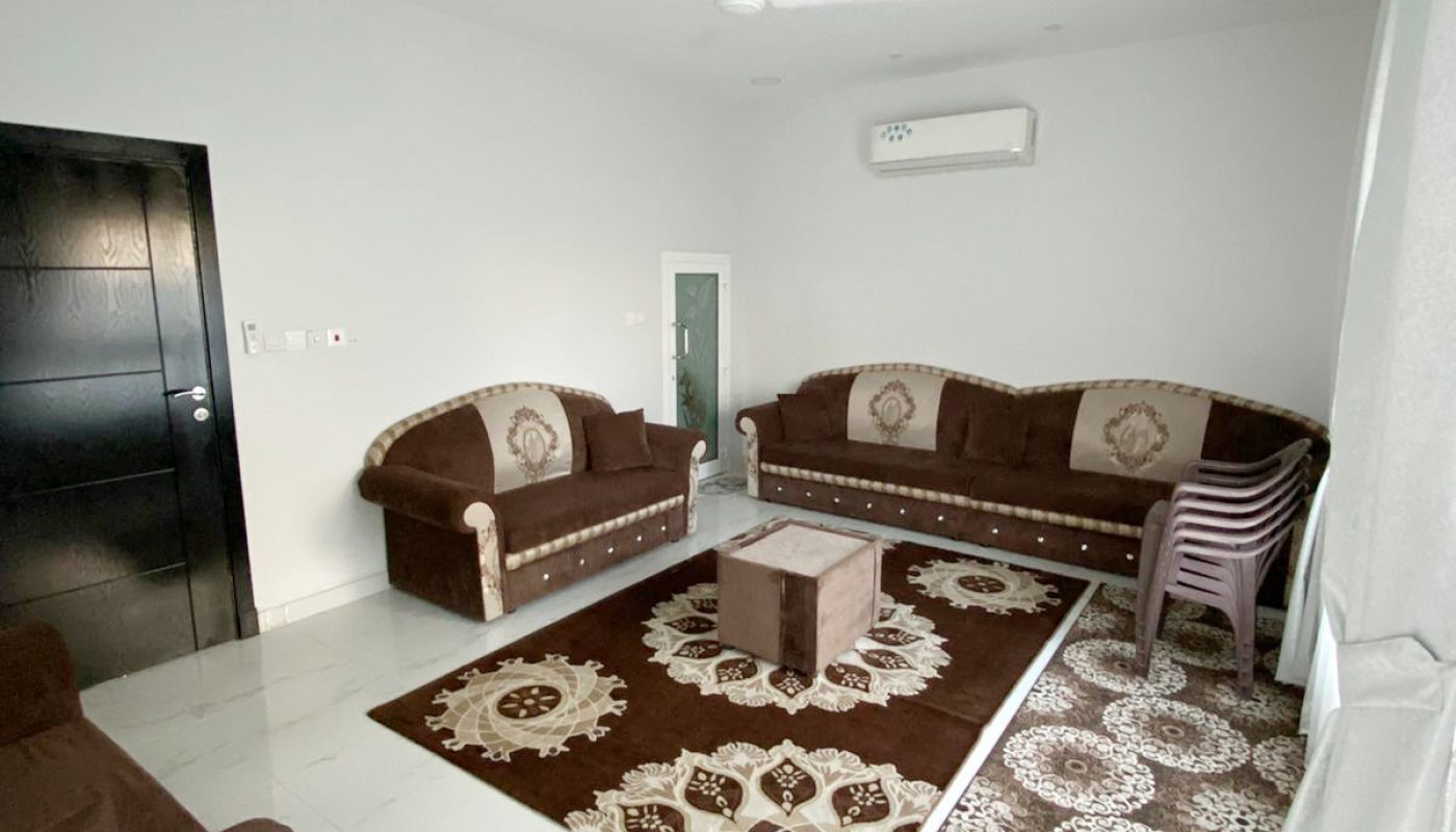 A living room with two brown sofas, a brown armchair, a central table, stacked plastic chairs, patterned rugs, a ceiling fan, and an air conditioner against white walls.