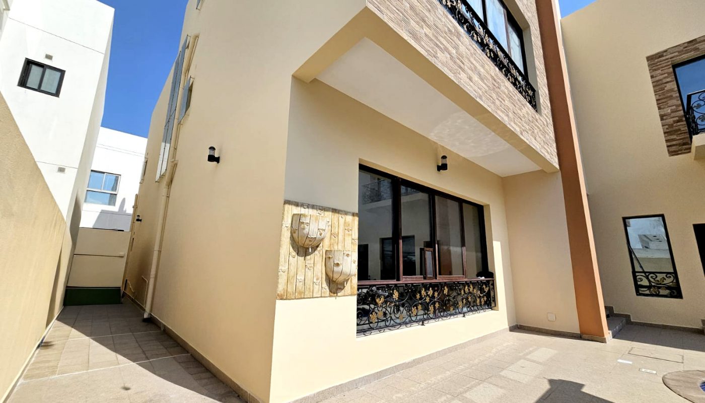 Modern two-story beige building with large windows, decorative metal railings, and outdoor wall lights, photographed on a sunny day with clear skies.