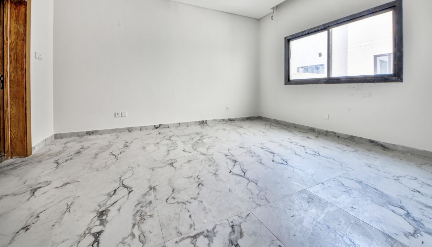 Empty room with marble tile flooring, white walls, a wooden door to the left, and a large rectangular window on the right wall.