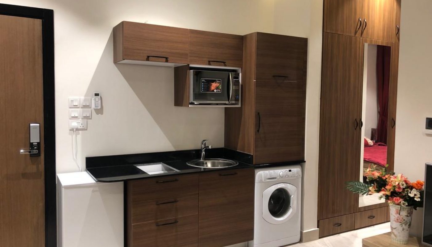 Compact modern kitchenette with wooden cabinets, a sink, washing machine, microwave, and wardrobe, adjacent to a door and a vase of flowers.