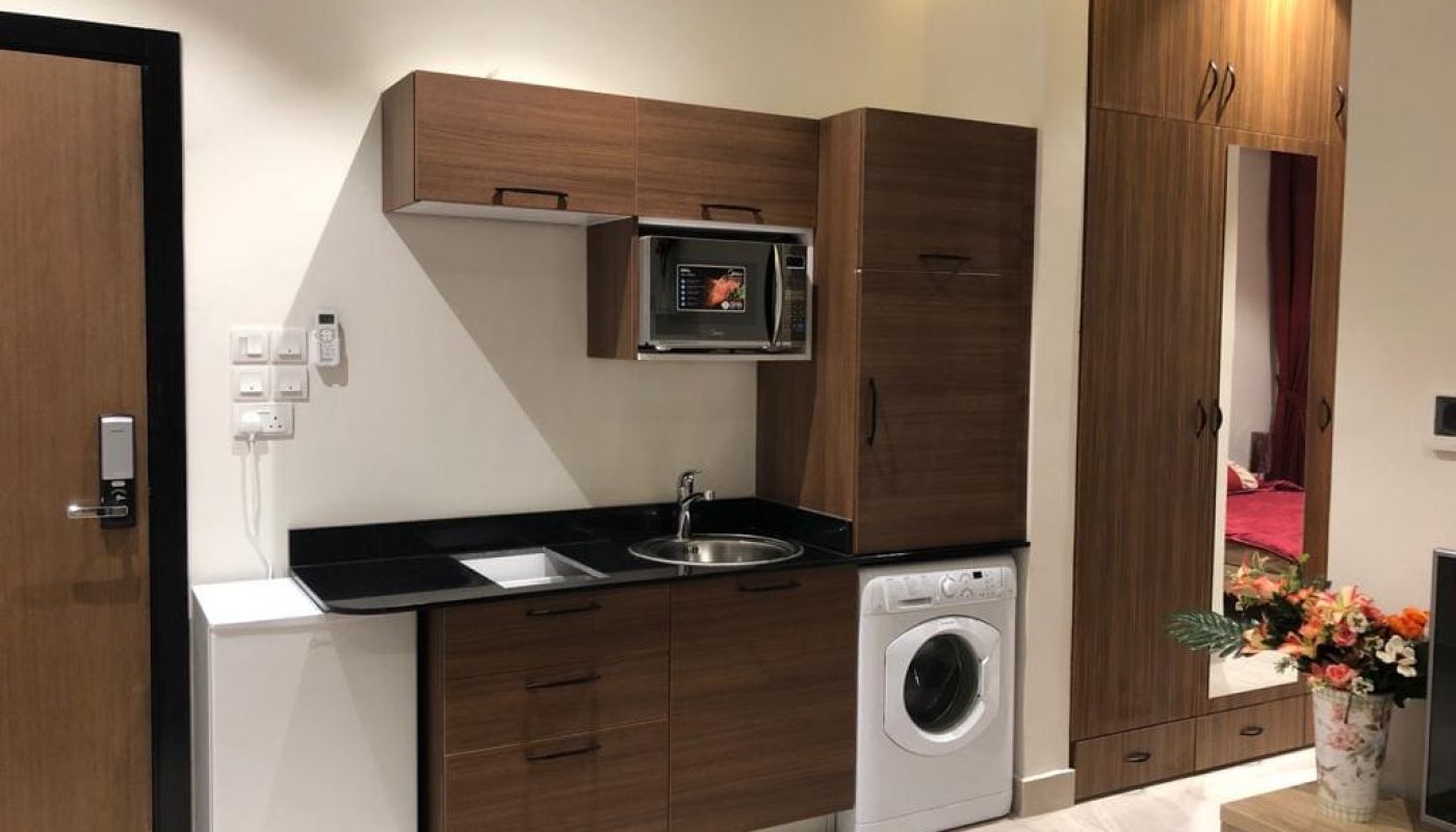 Compact modern kitchenette with wooden cabinets, a sink, washing machine, microwave, and wardrobe, adjacent to a door and a vase of flowers.