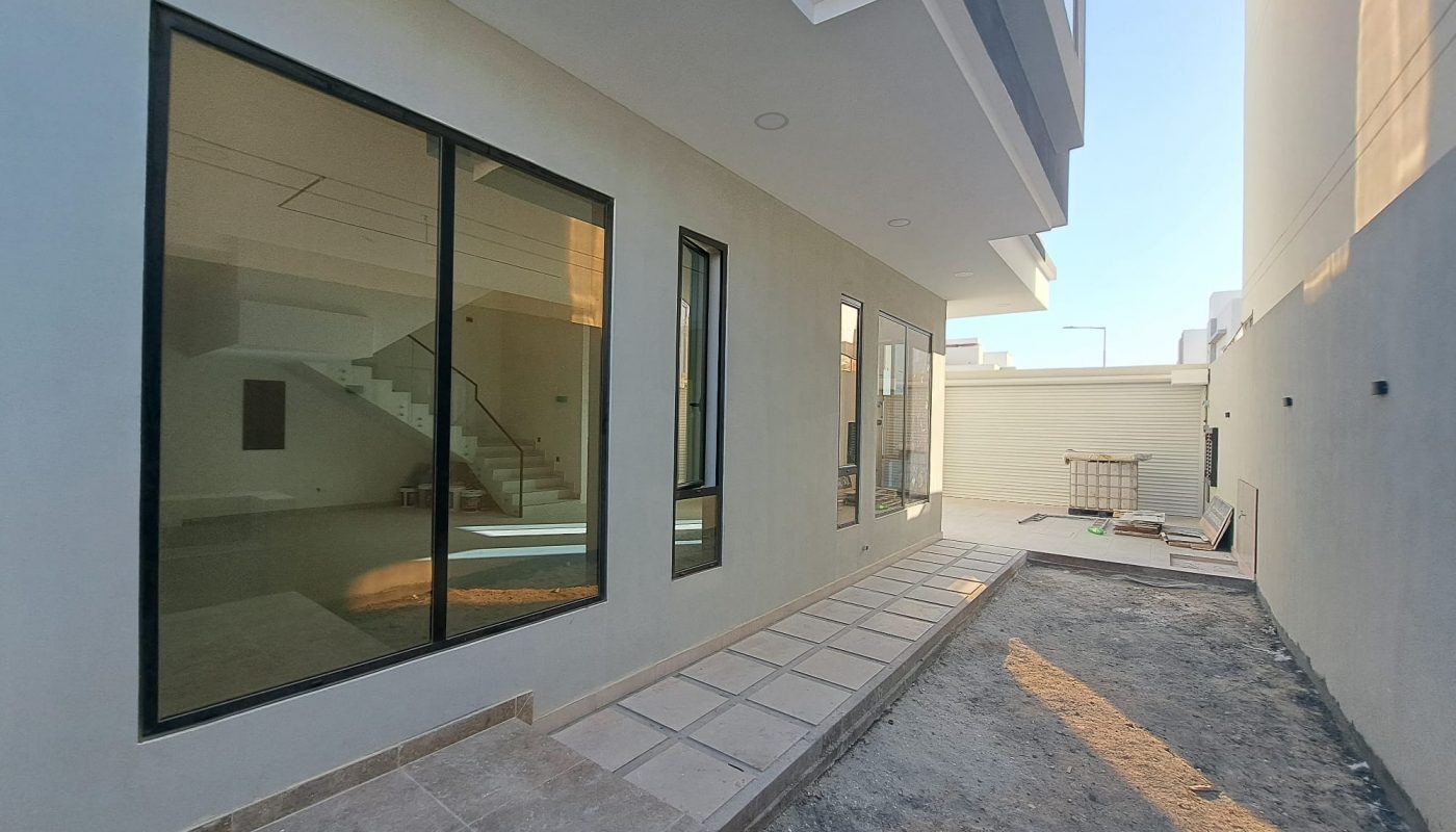 Exterior view of a modern house with large glass windows, a tiled walkway, and a dirt area beside the building under clear sky.