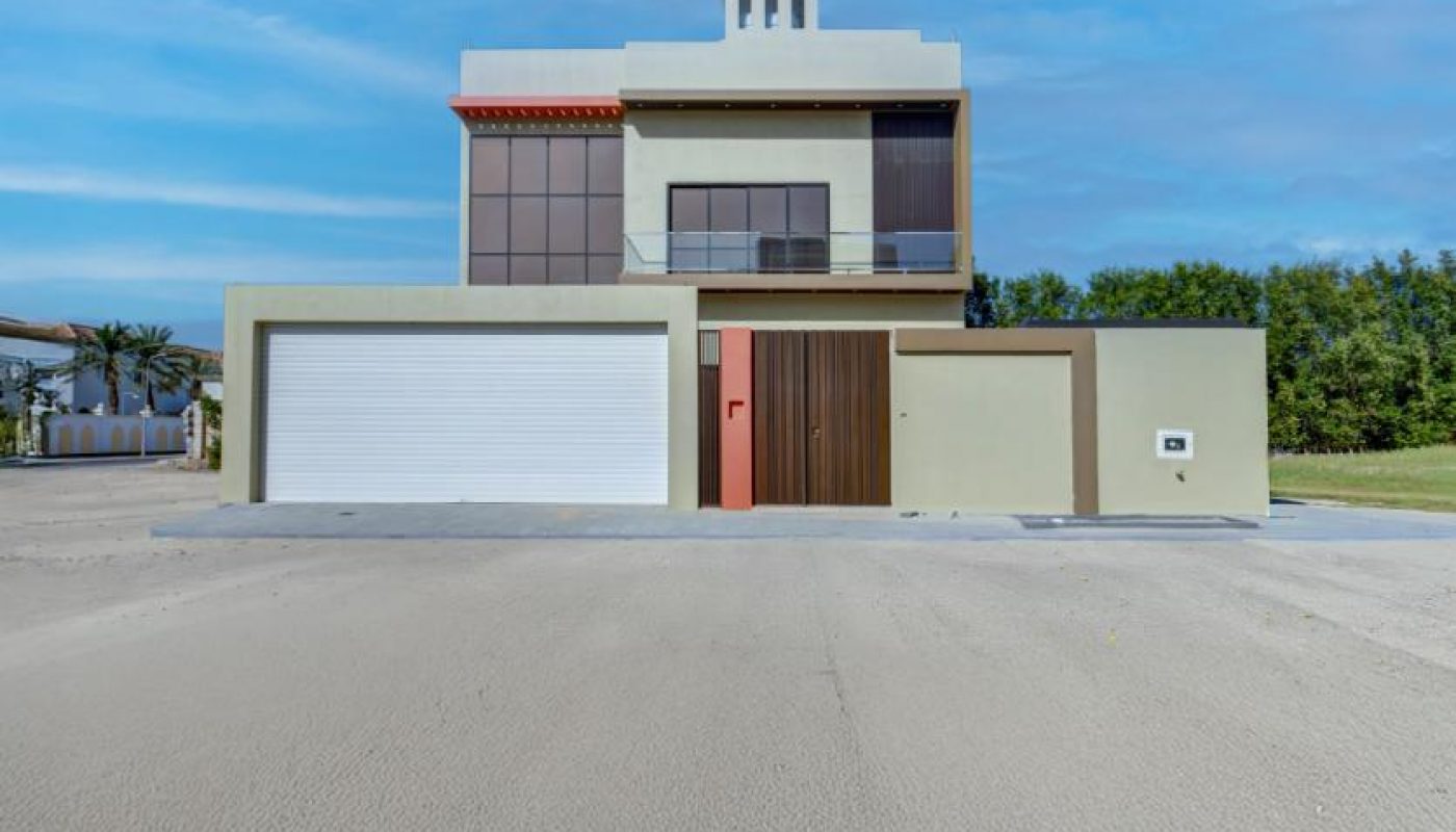Modern two-story house with large windows, a red front door, and a double garage, situated on a wide driveway under a clear blue sky.