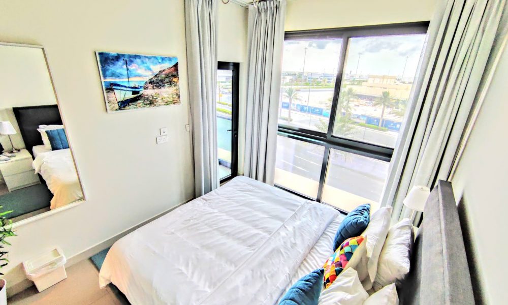 Bright bedroom with a large window, white bedding, blue pillows, a colorful cushion, a wall mirror, and artwork. A ceiling light fixture hangs above.