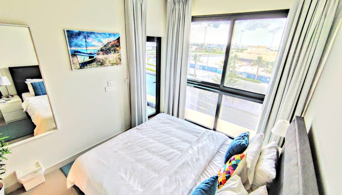 Bright bedroom with a large window, white bedding, blue accent pillows, wall art, curtains, and a ceiling light fixture.