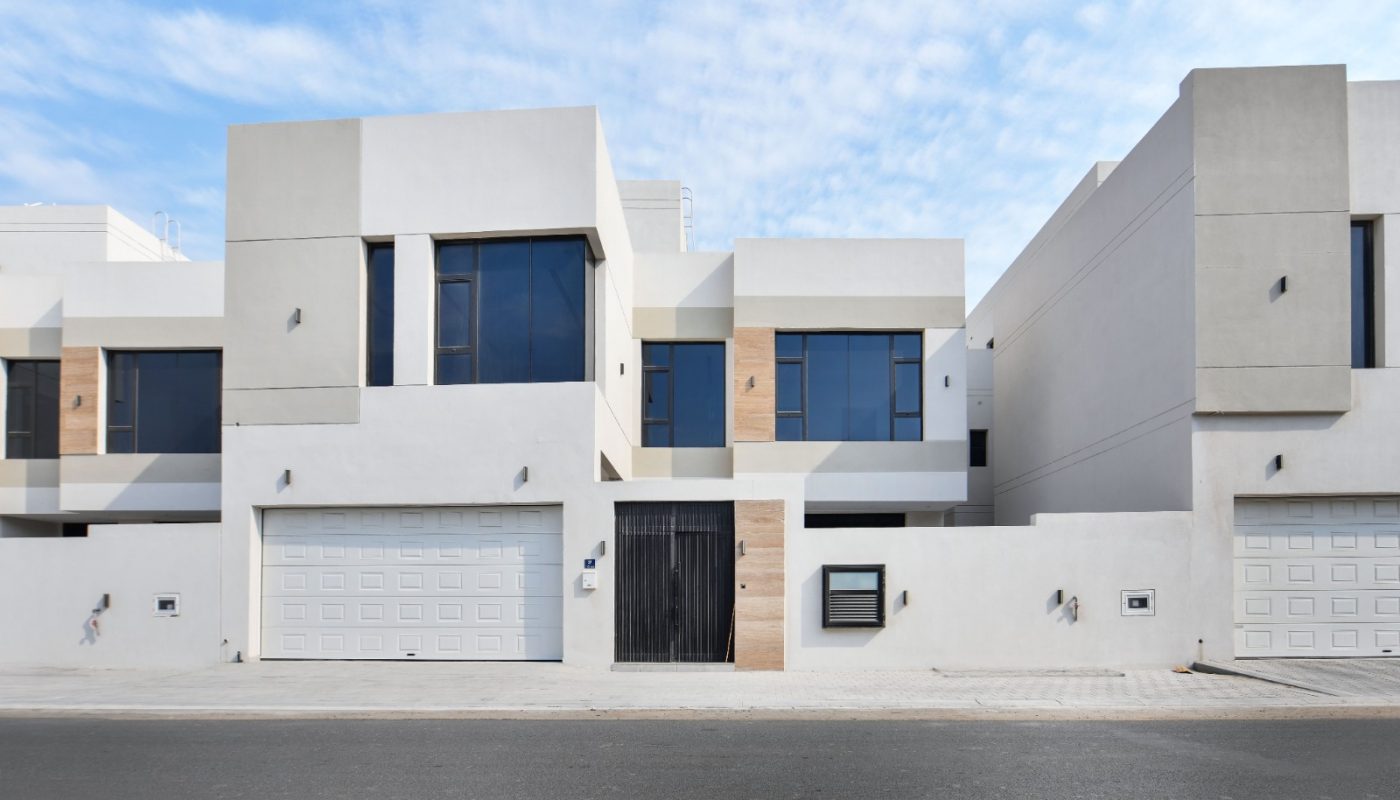 Modern two-story townhouse with large windows, white and beige exterior, two garages, and a central black gate facing a paved street.