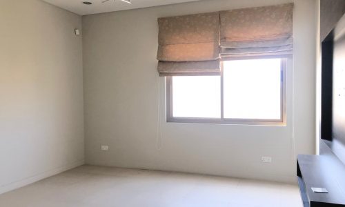 A minimalistic room with light-colored walls, a window with blinds, a ceiling light fixture, and a mounted TV unit. The room is empty and well-lit with natural light.