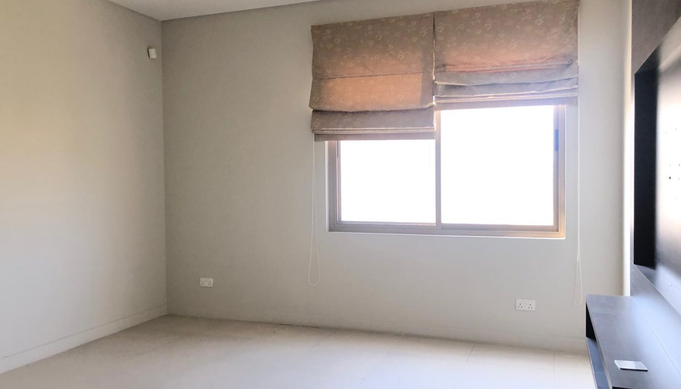 A minimalistic room with light-colored walls, a window with blinds, a ceiling light fixture, and a mounted TV unit. The room is empty and well-lit with natural light.