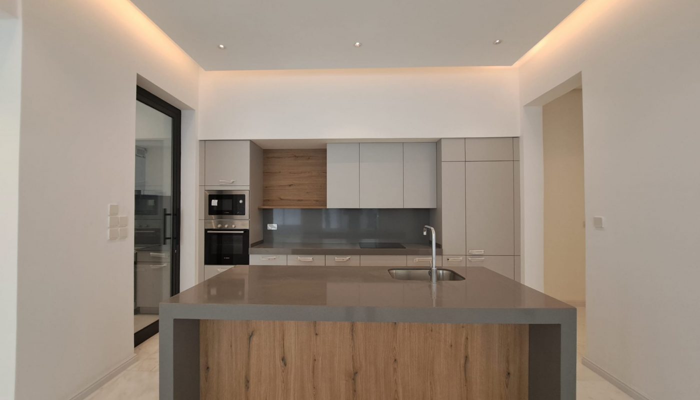 Modern kitchen with gray countertops, wood-accented island, built-in appliances, and under-cabinet lighting. Walls and floor are white, creating a clean, minimalist look.