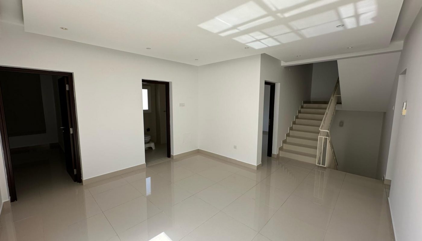 A bright, empty room with white walls, tiled floors, multiple doorways, and a staircase leading to an upper floor.