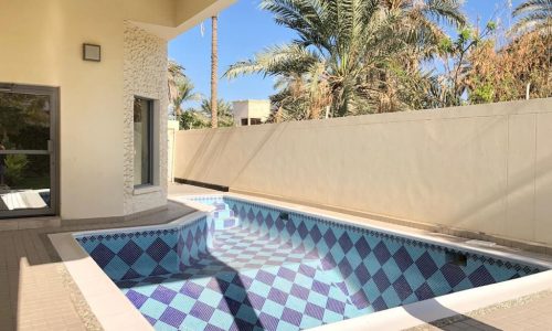 A small outdoor pool with blue and white tiles, surrounded by a patio and palm trees, adjacent to a light-colored building.