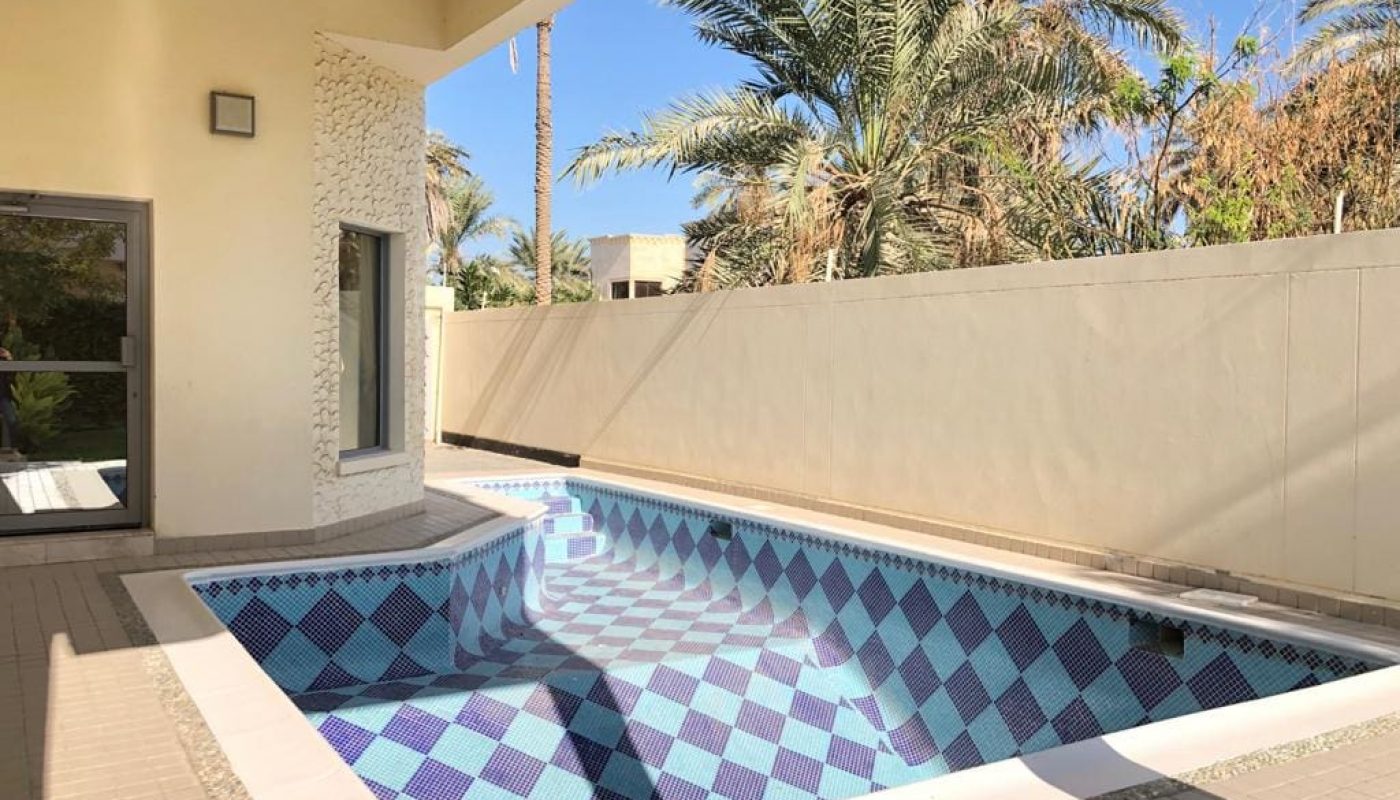 A small outdoor pool with blue and white tiles, surrounded by a patio and palm trees, adjacent to a light-colored building.