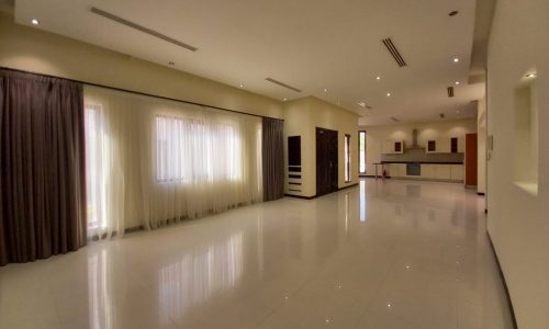 Spacious, empty living area with glossy tile flooring, large windows with curtains, and recessed ceiling lights, leading to an open kitchen at the far end.