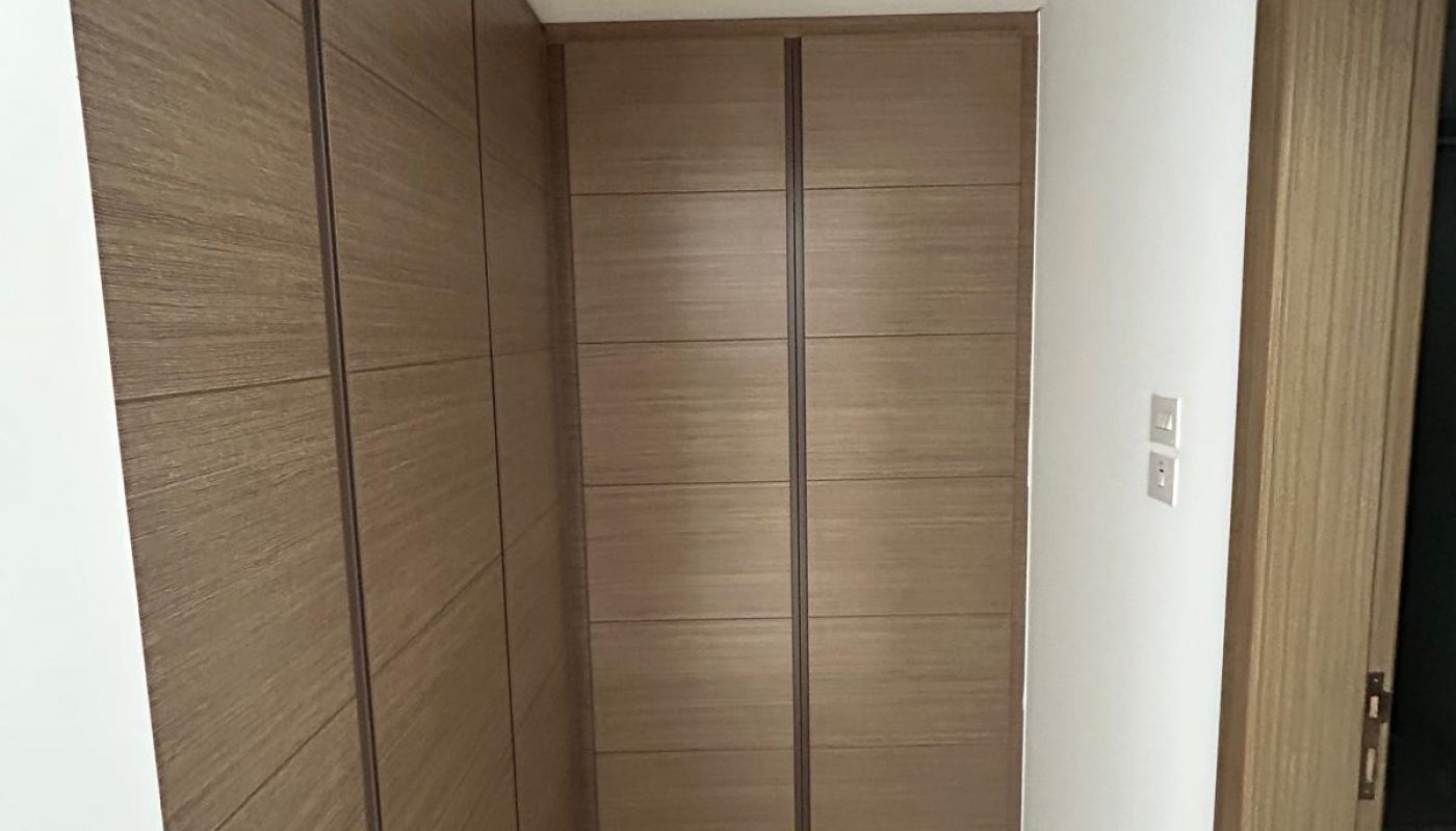 Small hallway with beige floor tiles, wooden wall panels on the left, and a large, closed wooden wardrobe at the end. White walls and a partially open wooden door on the right.