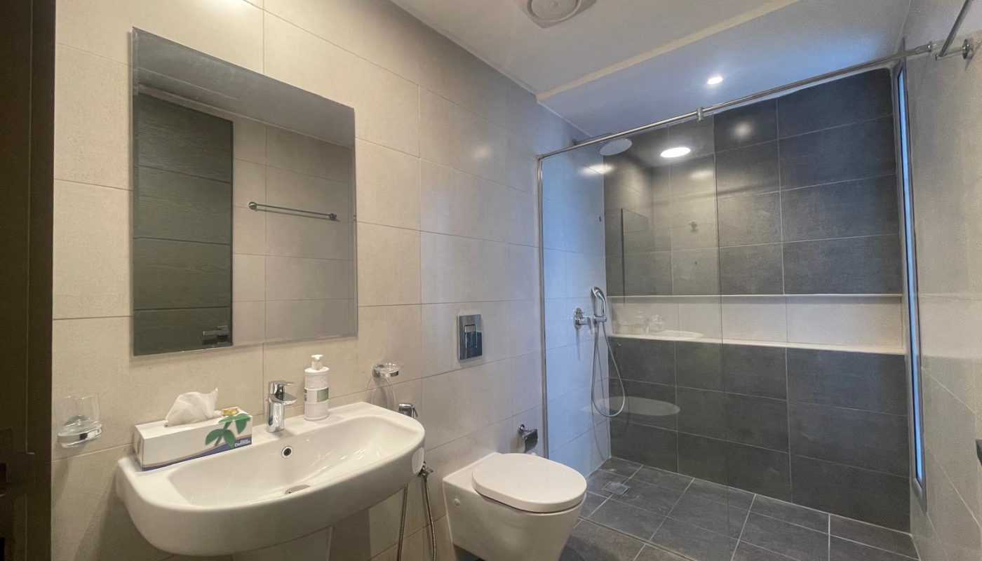 A modern bathroom with a wall-mounted sink, toilet, and bidet, large mirror, and a walk-in shower with glass partition and grey tile flooring and walls.