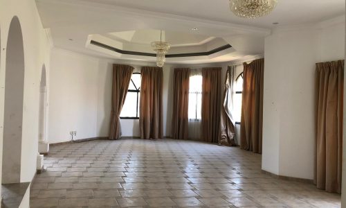 A spacious, empty room with tiled flooring, large windows with drawn beige curtains, and two ceiling chandeliers. The walls are white, and the room has a slightly elevated platform area on one side.