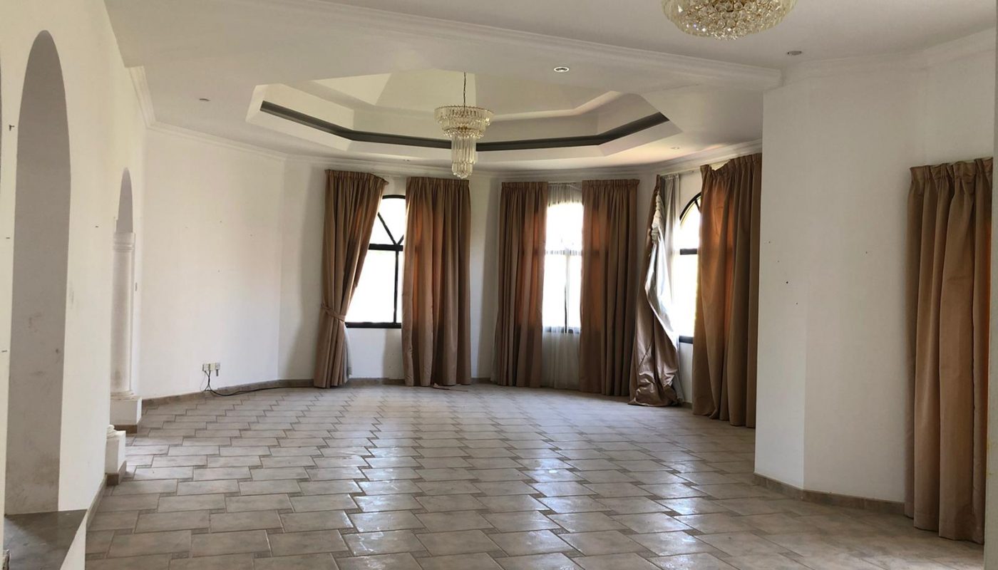 A spacious, empty room with tiled flooring, large windows with drawn beige curtains, and two ceiling chandeliers. The walls are white, and the room has a slightly elevated platform area on one side.