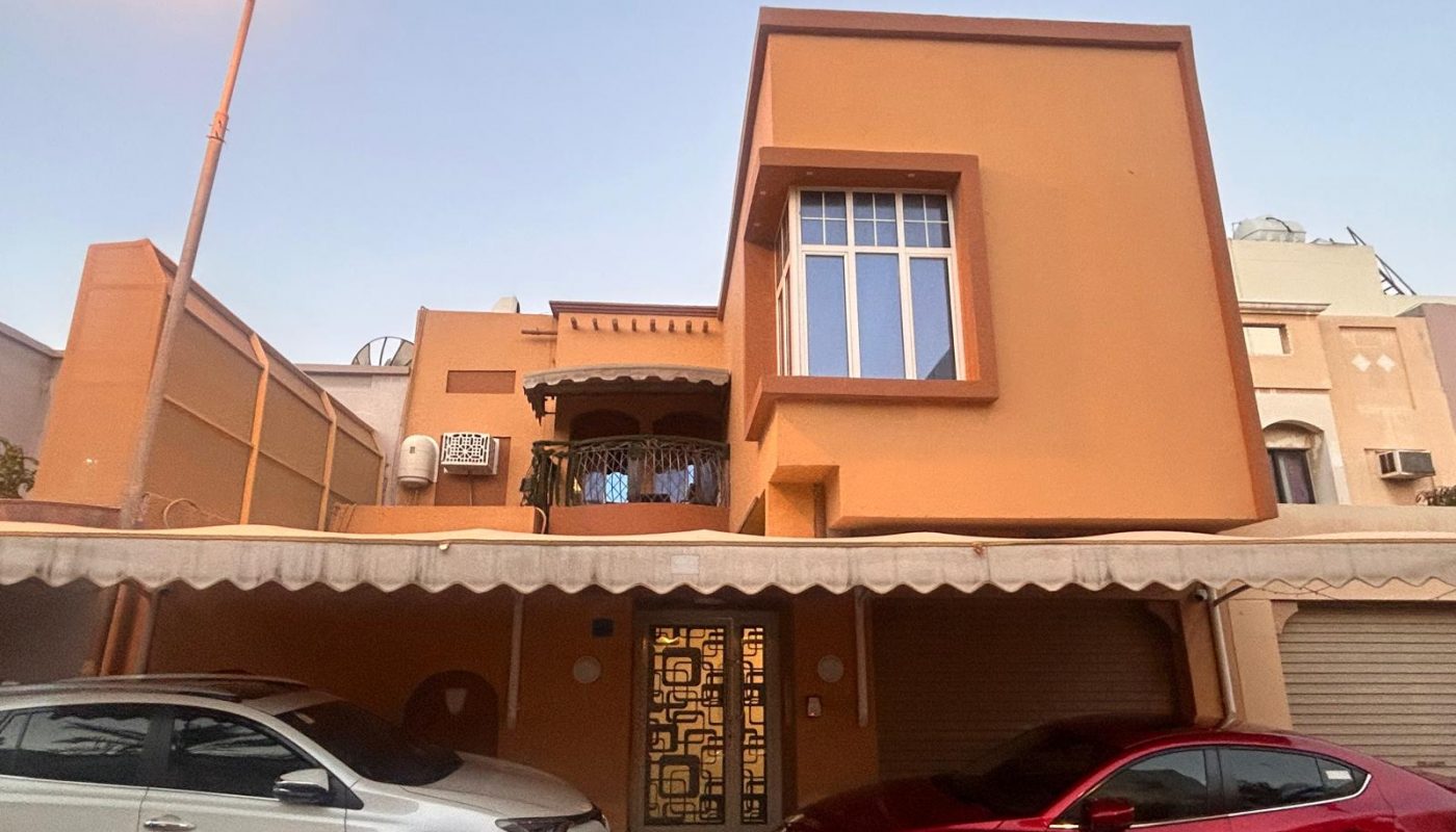 Two cars parked in front of a two-story, orange house with a balcony, large window, gated entrance, and a streetlamp on the left.