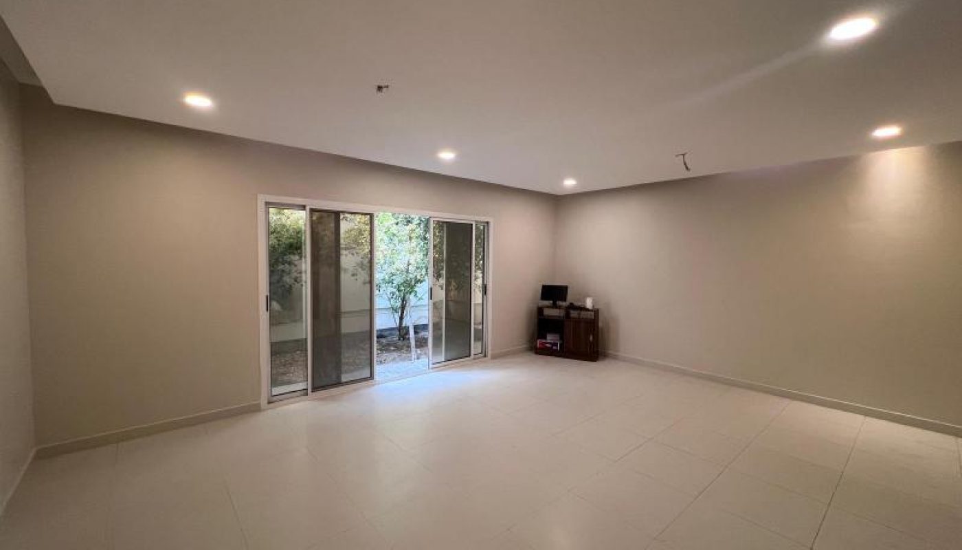 A spacious, empty room with tiled flooring, recessed ceiling lights, sliding glass doors, and a small desk in the corner near the wall.
