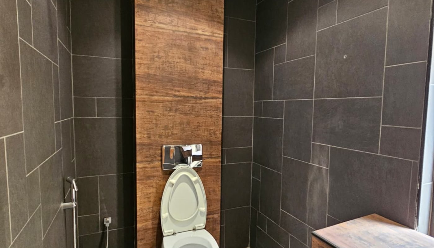 A modern bathroom with dark tiles, featuring a wall-mounted toilet with the lid open, a handheld bidet spray, and a wooden accent wall.