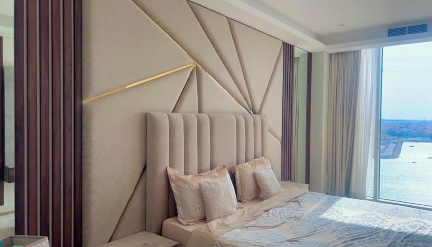 A modern bedroom with a tan upholstered headboard, geometric wall design, and large window with a view of a body of water and distant cityscape.