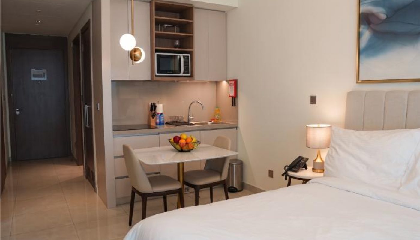 Modern hotel room with a neatly made bed, bedside lamp, small kitchenette, dining table with two chairs, and a bowl of fruit. Doorway and hallway visible in the background.