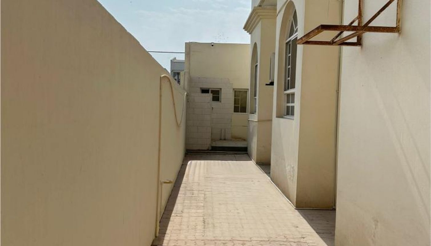 A narrow paved walkway runs alongside a cream-colored building with two windows, ending at a small structure in the background under a clear sky.