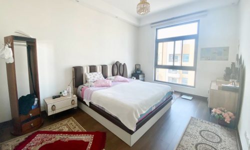 A bedroom with a large bed, patterned rugs, a small desk, and a window. Clothing hangs on a garment rack, and a chandelier is on the ceiling.