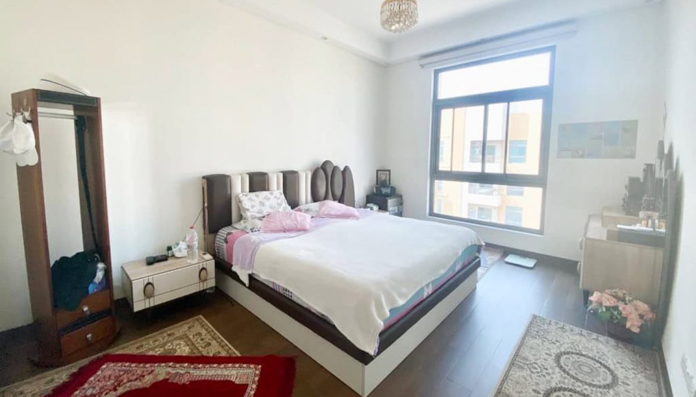 A bedroom with a large bed, patterned rugs, a small desk, and a window. Clothing hangs on a garment rack, and a chandelier is on the ceiling.