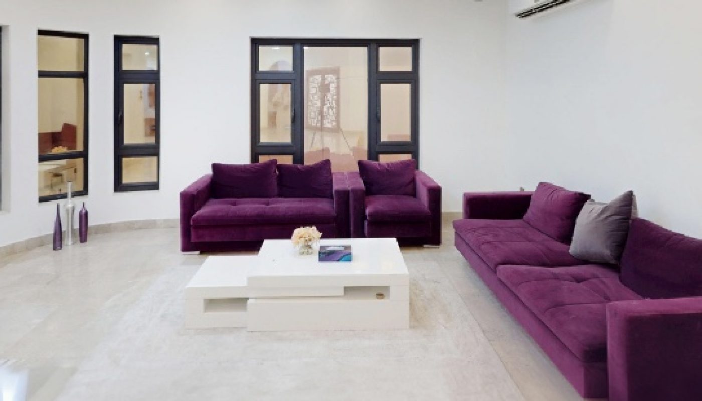 A modern living room with purple sofas, a white coffee table, a light rug, and multiple windows. The ceiling features recessed lighting and yellow accents.