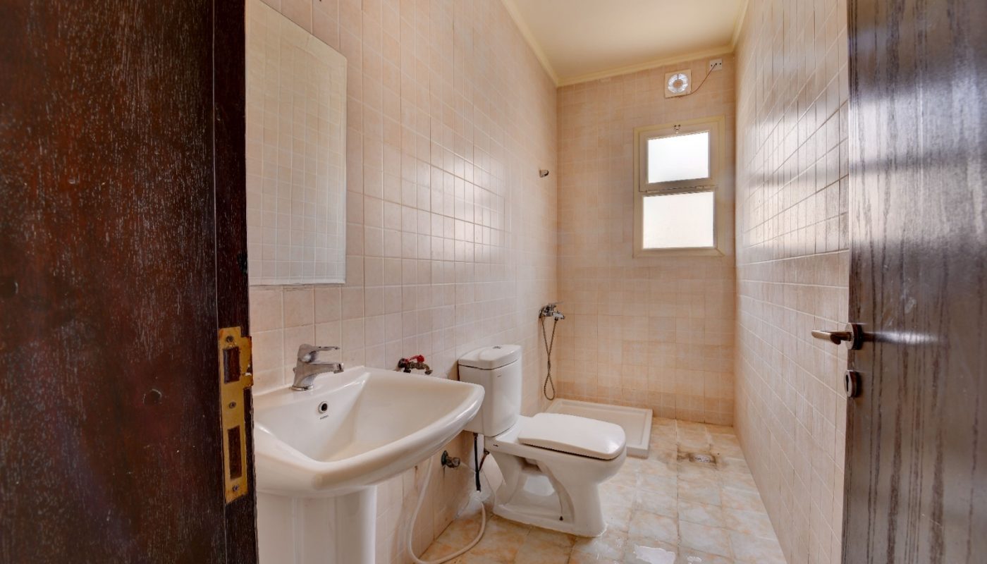 A simple bathroom with a sink, mirror, toilet, bidet shower, and a window, featuring beige tiles on the walls and floor.