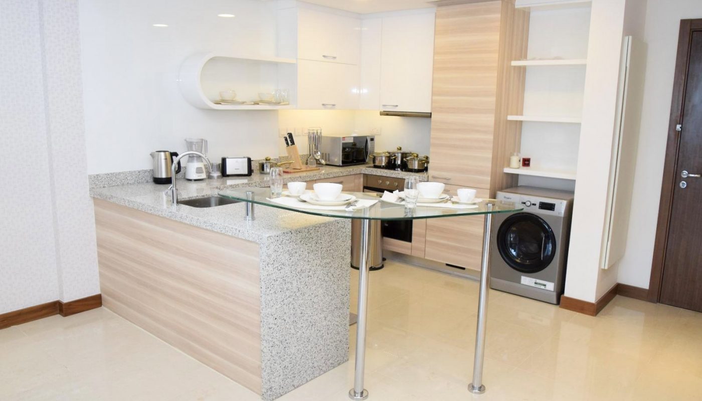 Modern kitchen with light wood cabinets, granite countertops, a built-in washing machine, appliances, and a glass breakfast bar set with cups and saucers.