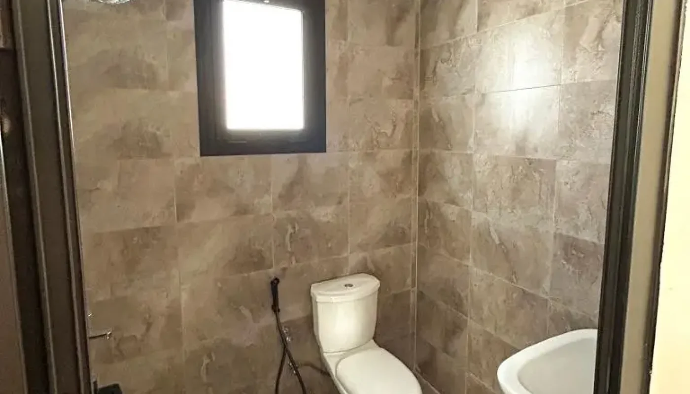 A simple bathroom with a toilet, handheld bidet, wall-mounted sink, window, and brown tiled walls and floor.