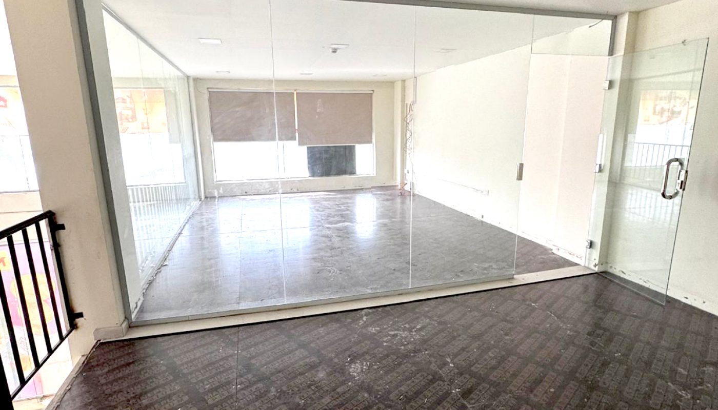 A glass-walled room with a closed glass door on one side, empty interior, tiled floors, and a large window with a brown blind in the background.