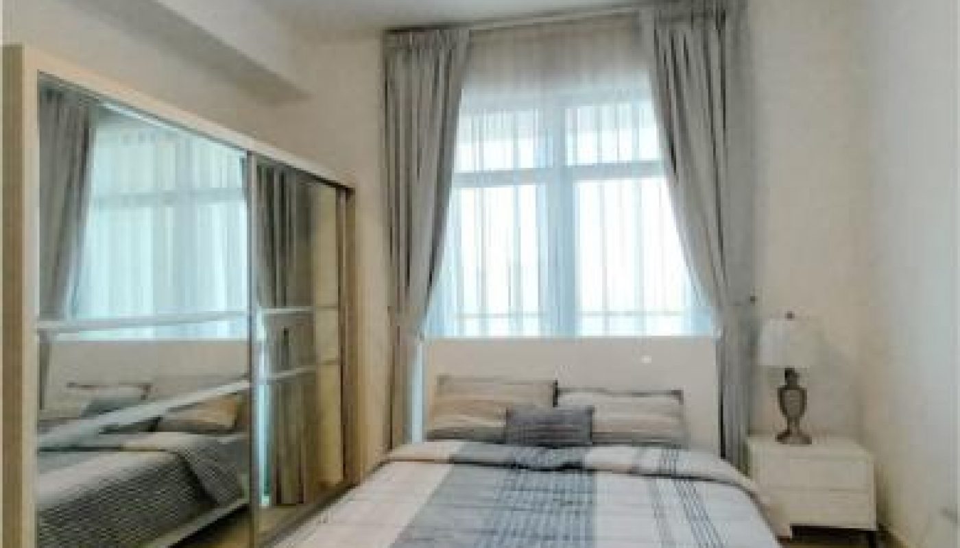 A small bedroom with a double bed, plaid bedding, a mirrored wardrobe, a window with curtains, and a bedside table with a lamp.