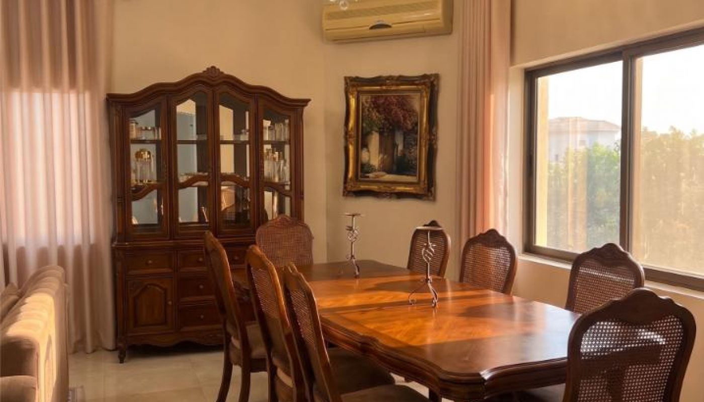 A dining room with a wooden table, six chairs, a china cabinet, chandelier, and large windows with curtains. A framed painting hangs on the wall above the air conditioner.