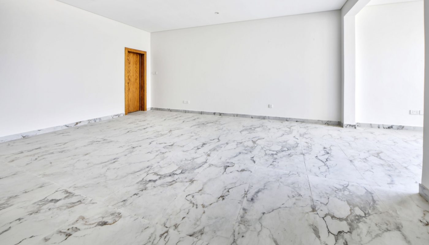 Empty room with white walls, large marble floor tiles, a wooden door, and a wide open doorway leading to another space. Ceiling has a small red fire alarm fixture.