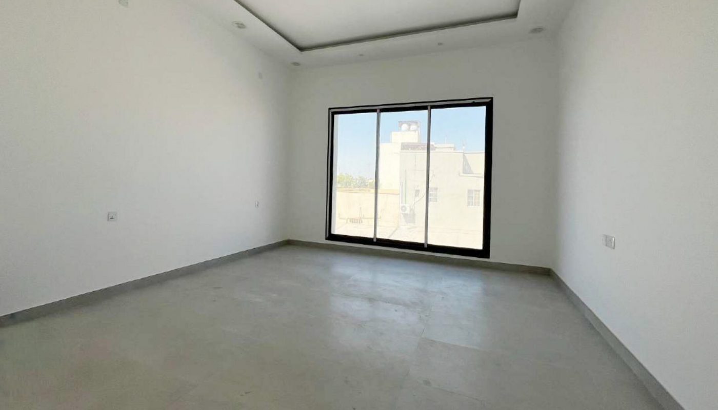 Empty, unfurnished room with white walls, gray tiled floor, recessed lighting, and a large window overlooking buildings outside.