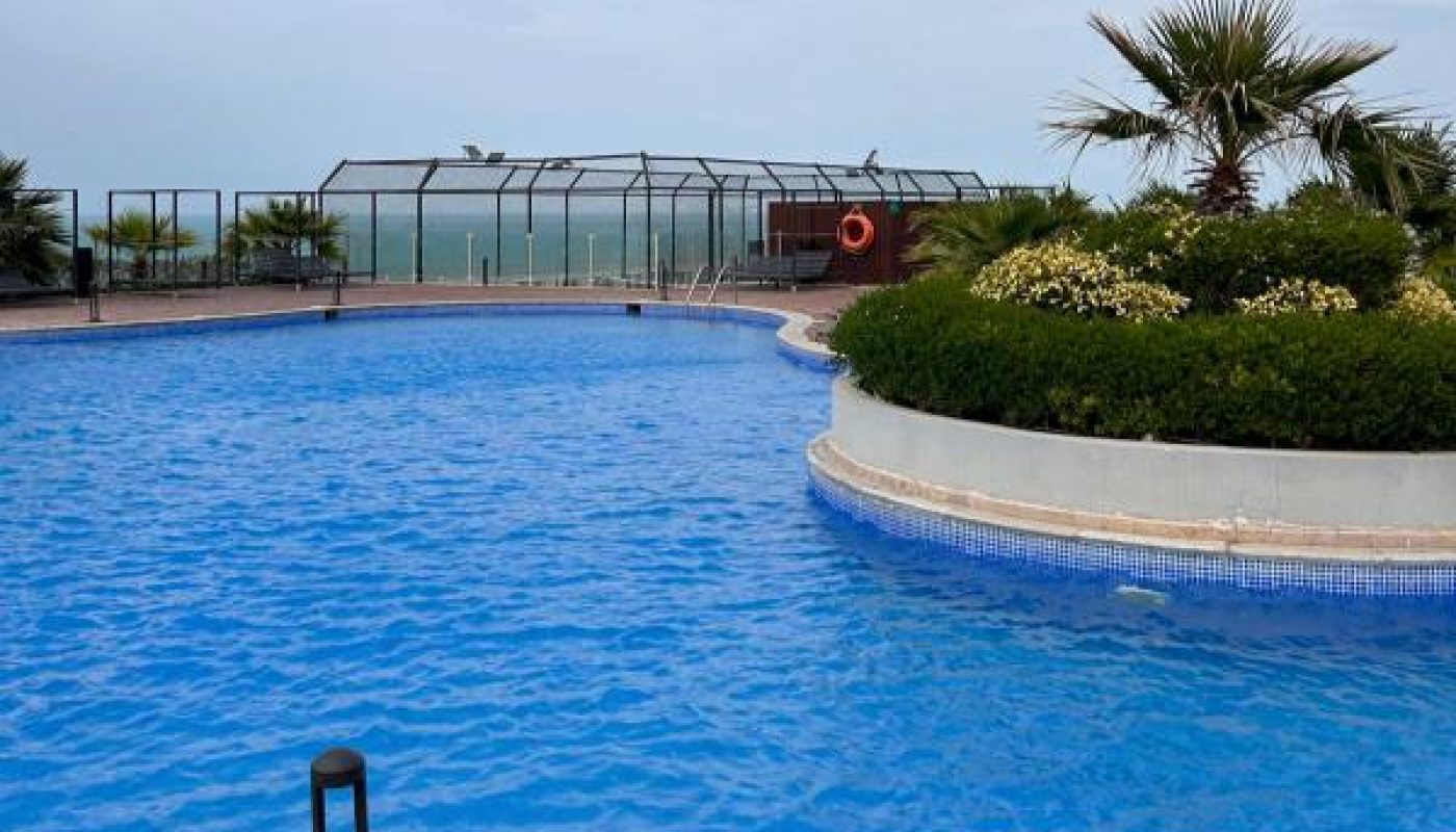 Outdoor swimming pool with clear blue water, wooden deck marked "2M", plants nearby, and a fenced area overlooking the ocean under a cloudy sky.